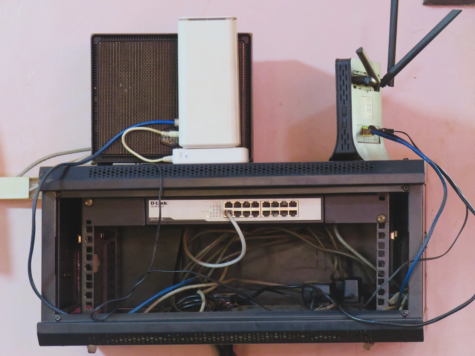 Duvara monte ağ rafında Ethernet anahtarı ve kablolar, rafın üzerinde antenli yönlendirici ve beyaz cihazlar.